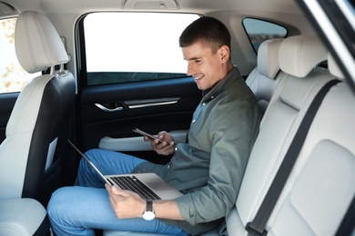Photo of Attractive young man working with laptop and smartphone on backseat in luxury car