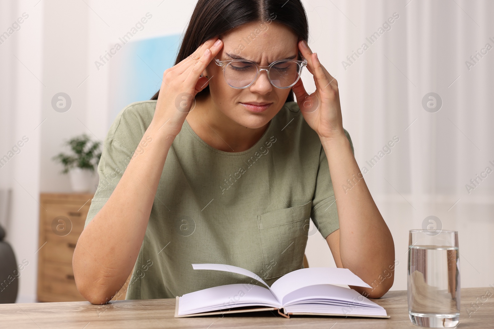 Photo of Sad woman suffering from headache at wooden table with open book and glass of water indoors