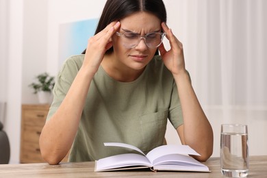 Photo of Sad woman suffering from headache at wooden table with open book and glass of water indoors