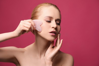 Photo of Beautiful young woman doing facial massage with gua sha tool on pink background