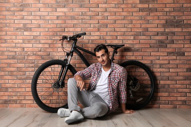 Photo of Handsome young man with modern bicycle near brick wall indoors