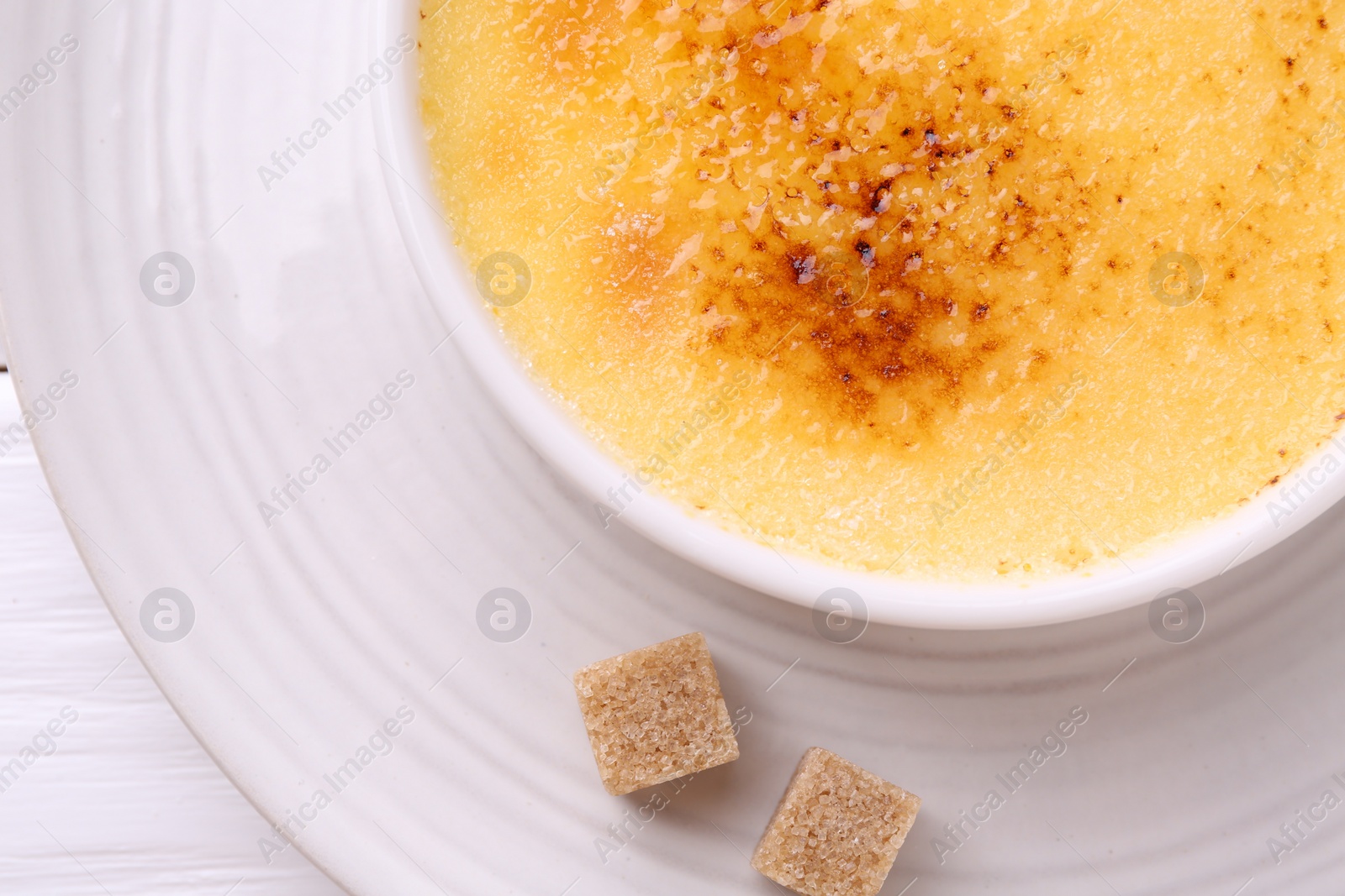 Photo of Delicious creme brulee in bowl and sugar cubes on white wooden table, top view