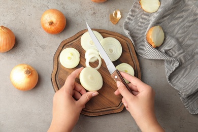 Photo of Young woman cutting ripe onion on wooden board at grey table, top view