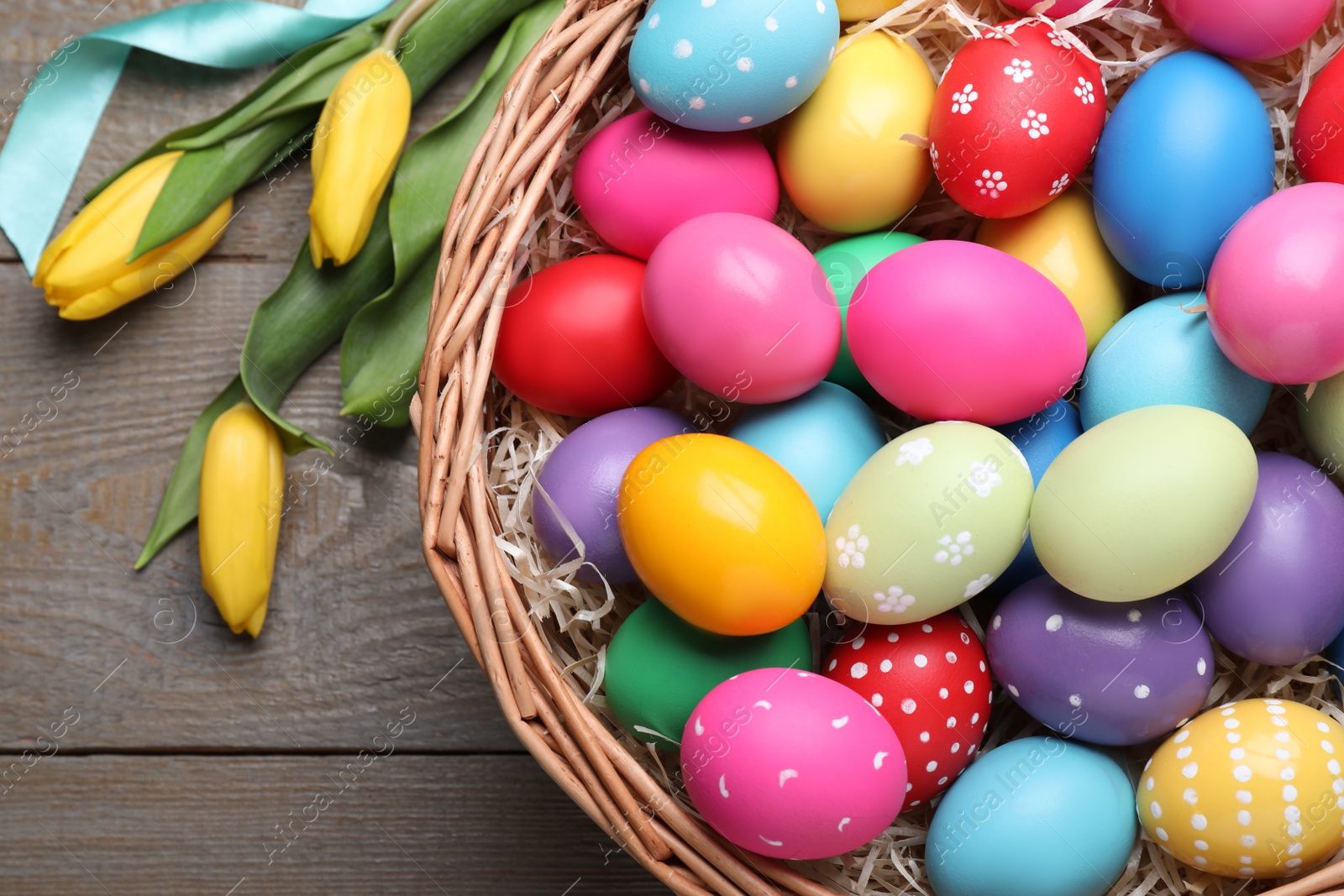 Photo of Colorful Easter eggs in wicker basket and tulips on wooden table, flat lay