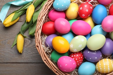 Photo of Colorful Easter eggs in wicker basket and tulips on wooden table, flat lay