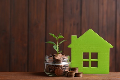 Photo of House model, jar with coins and plant on table against wooden background. Space for text
