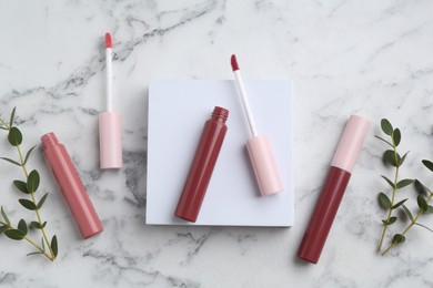 Photo of Different lip glosses, applicators, green leaves and podium on white marble table, flat lay