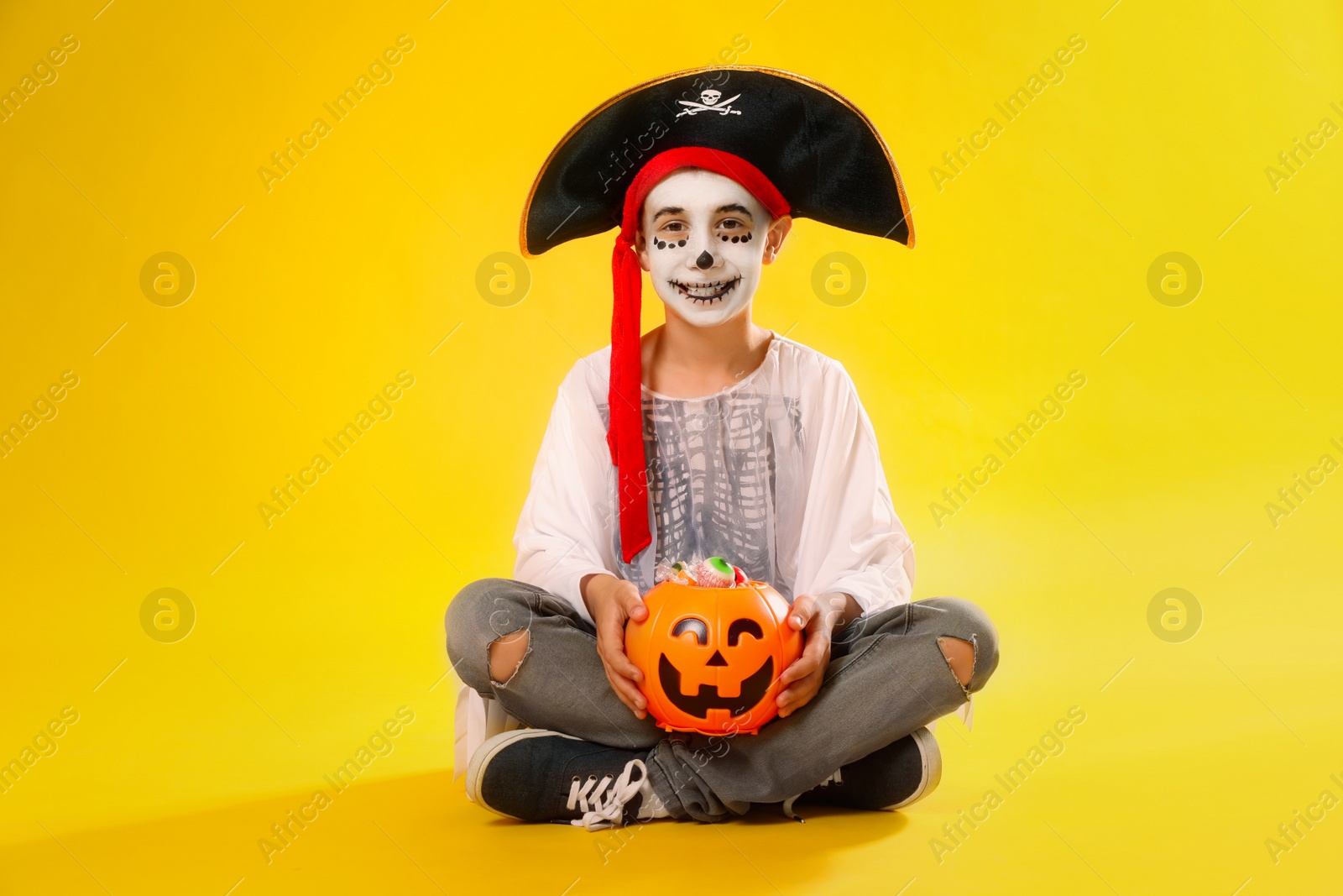 Photo of Cute little boy with pumpkin candy bucket wearing Halloween costume on yellow background