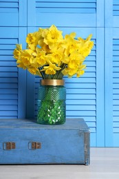 Vase with beautiful daffodils on wooden crate near light blue folding screen indoors
