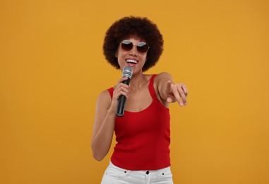 Curly young woman in sunglasses with microphone singing on yellow background