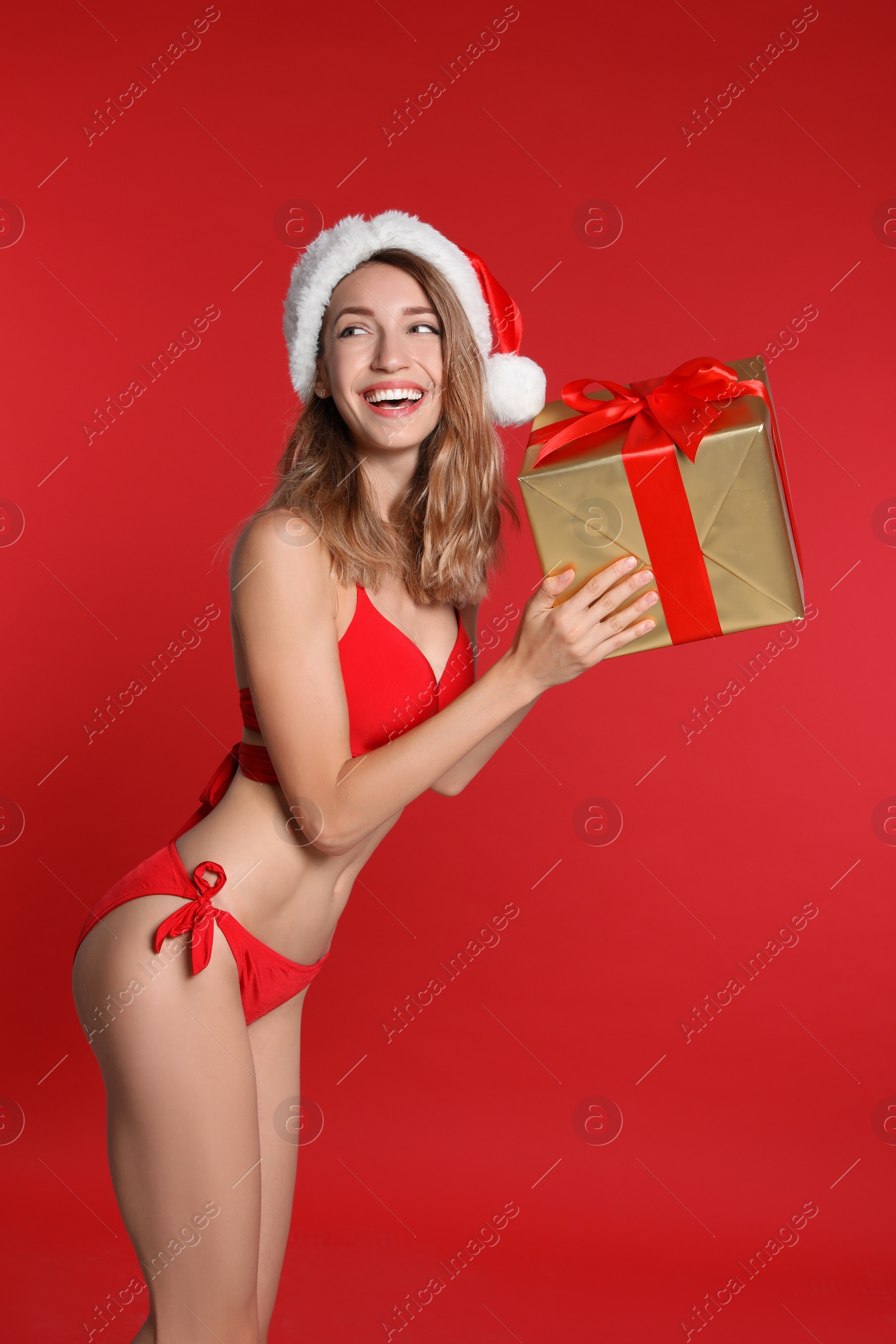 Photo of Sexy young woman in bikini and Santa hat with gift box on red background. Christmas celebration