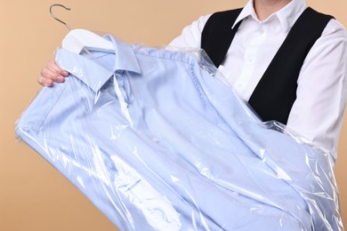 Photo of Dry-cleaning service. Woman holding shirt in plastic bag on beige background, closeup