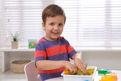 Cute little boy playing with bright kinetic sand at table in room