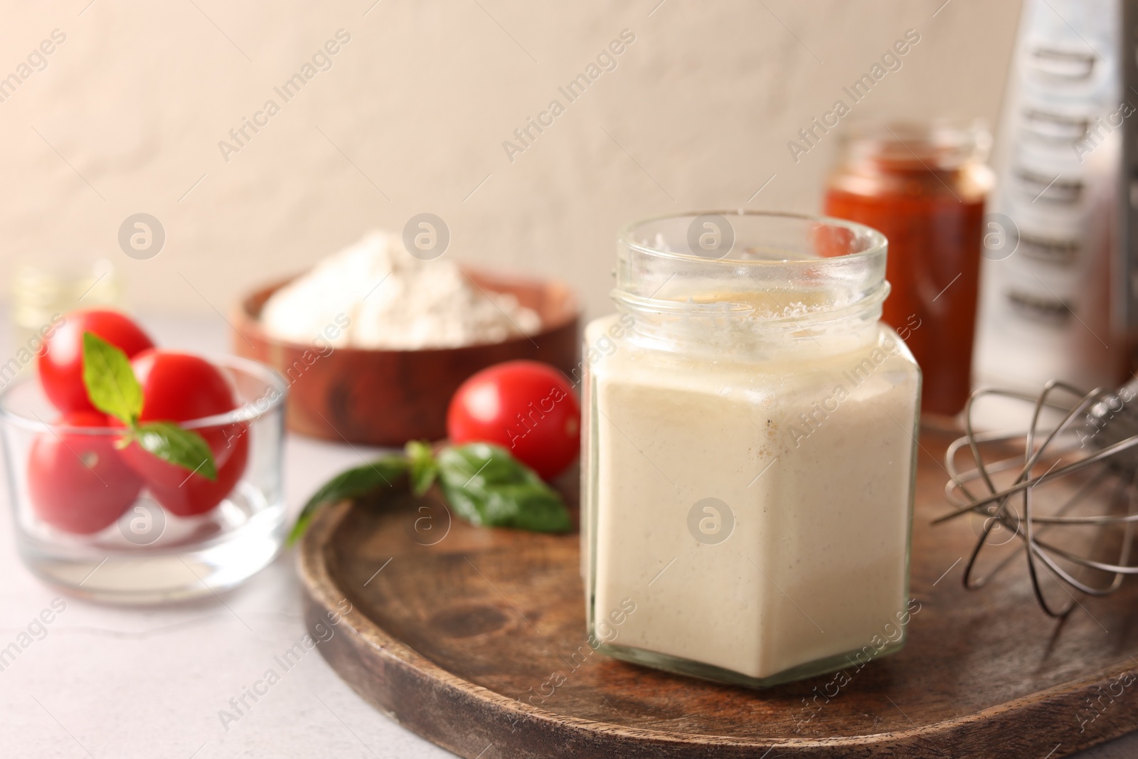 Photo of Pizza dough starter in glass jar, products and whisk on gray table