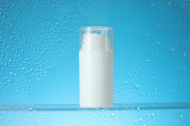 Photo of Bottle with moisturizing cream on light blue background, view through wet glass