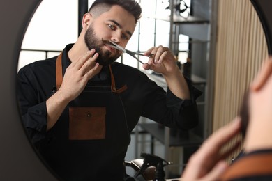 Photo of Handsome young man trimming mustache with scissors near mirror indoors