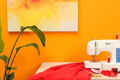 Modern sewing machine with cloth and craft accessories on wooden table near orange wall