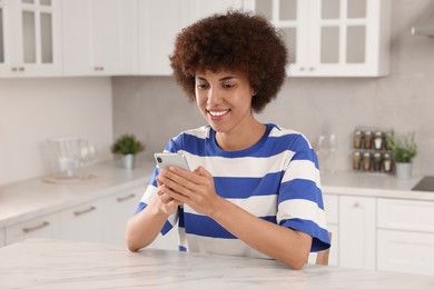 Happy young woman with smartphone at table in kitchen
