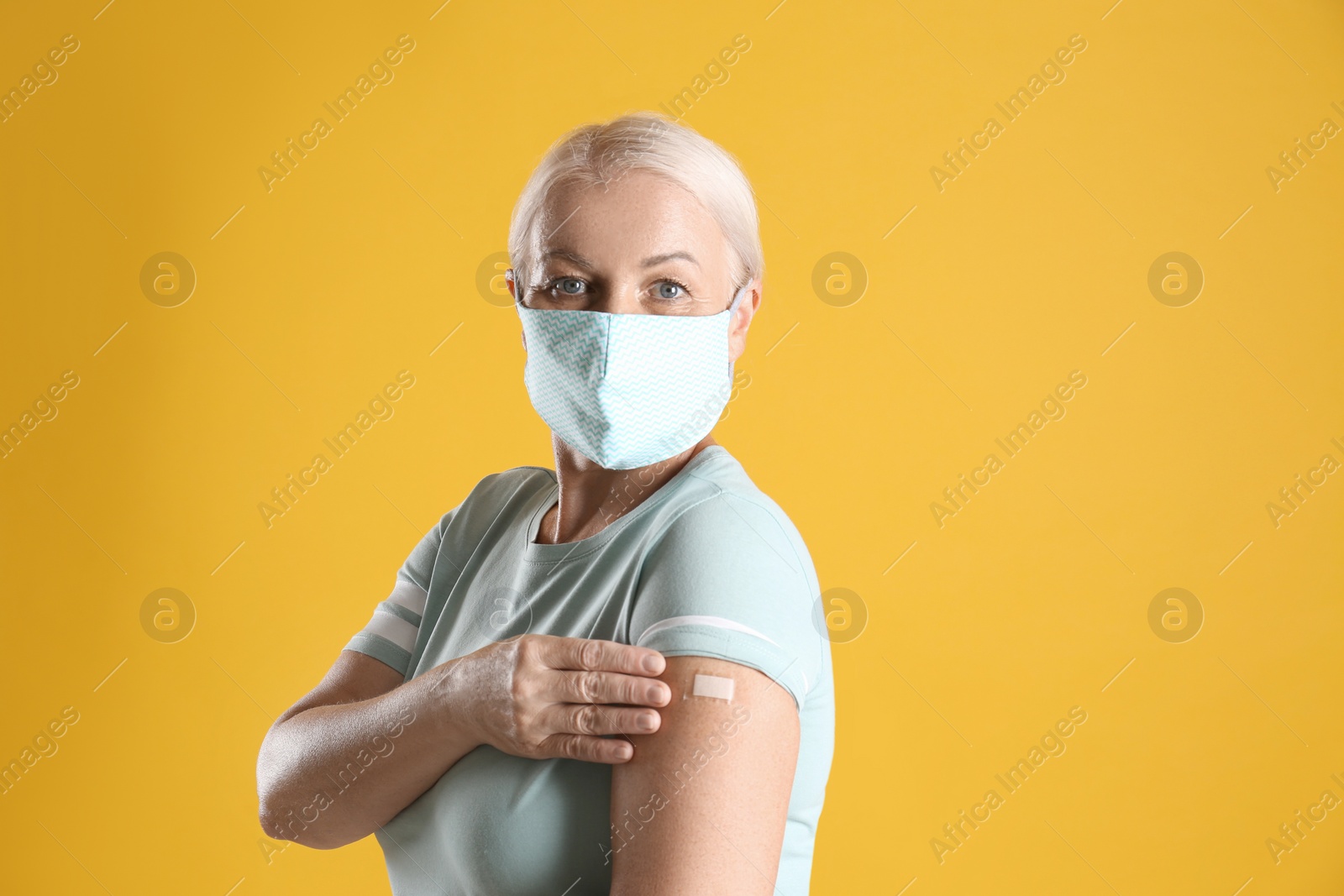 Photo of Mature woman in protective mask showing arm with bandage after vaccination on yellow background