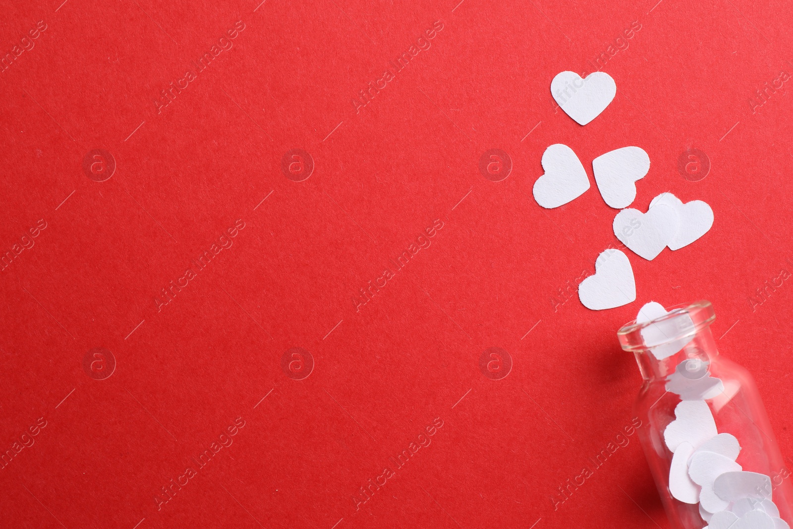 Photo of White paper hearts scattered from small glass bottle on red background, flat lay. Space for text