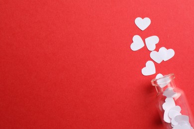 Photo of White paper hearts scattered from small glass bottle on red background, flat lay. Space for text