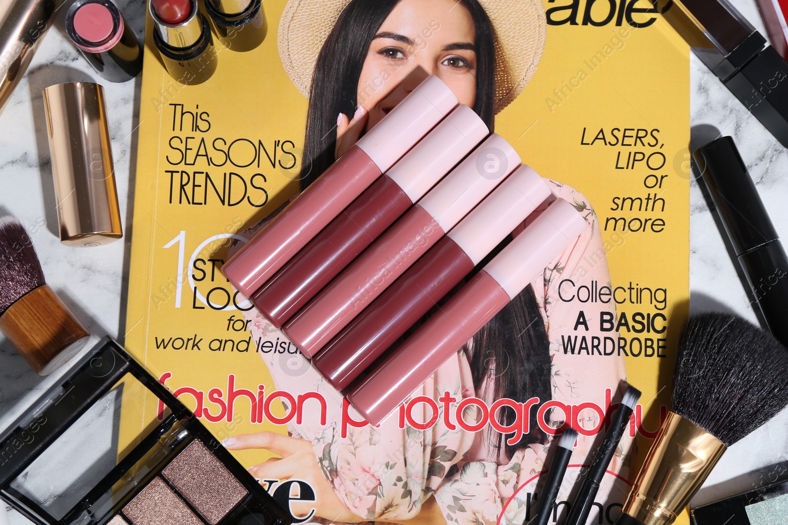 Photo of Bright lip glosses among different cosmetic products and fashion magazine on table, flat lay