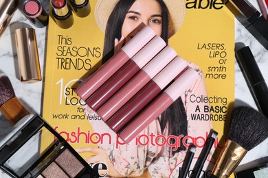 Bright lip glosses among different cosmetic products and fashion magazine on table, flat lay