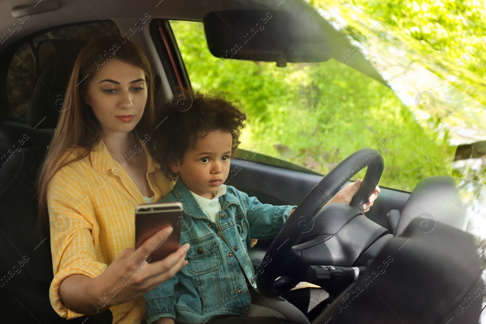 Photo of Mother with little daughter driving car and chatting on phone. Child in danger