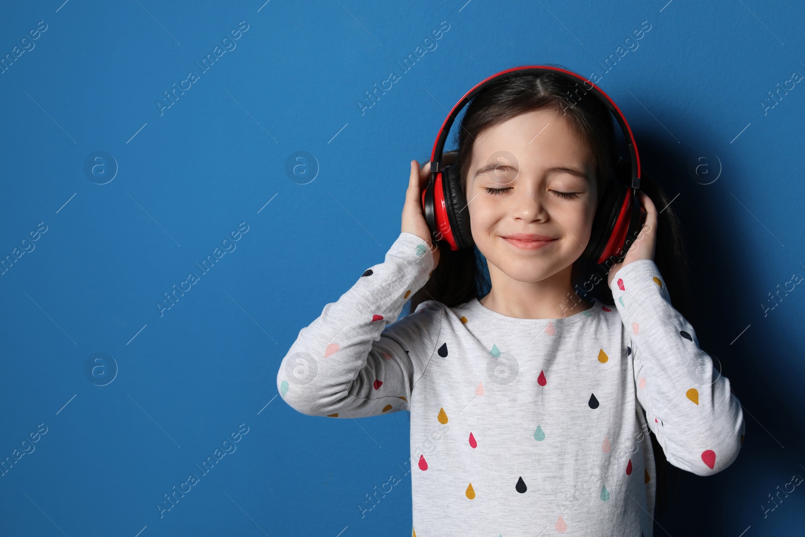 Photo of Cute little girl with headphones listening to audiobook on blue background. Space for text