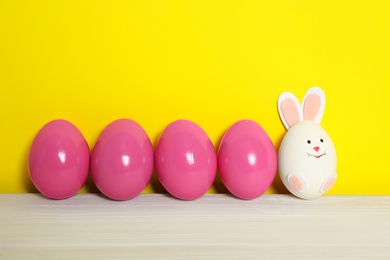 Photo of Bright Easter eggs and white one as cute bunny on wooden table against yellow background