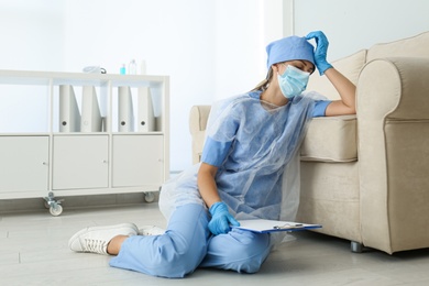 Photo of Exhausted doctor sitting on floor indoors. Stress of health care workers during COVID-19 pandemic