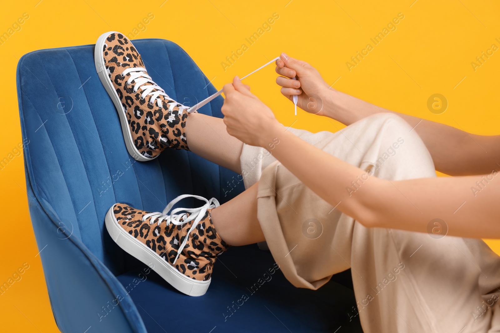 Photo of Woman lacing up classic old school sneakers with leopard print on armchair against orange background, closeup