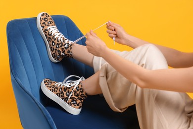 Photo of Woman lacing up classic old school sneakers with leopard print on armchair against orange background, closeup