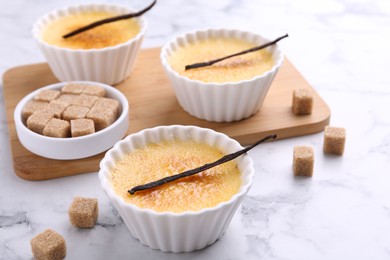 Delicious creme brulee in bowls, vanilla pods and sugar cubes on white marble table, closeup