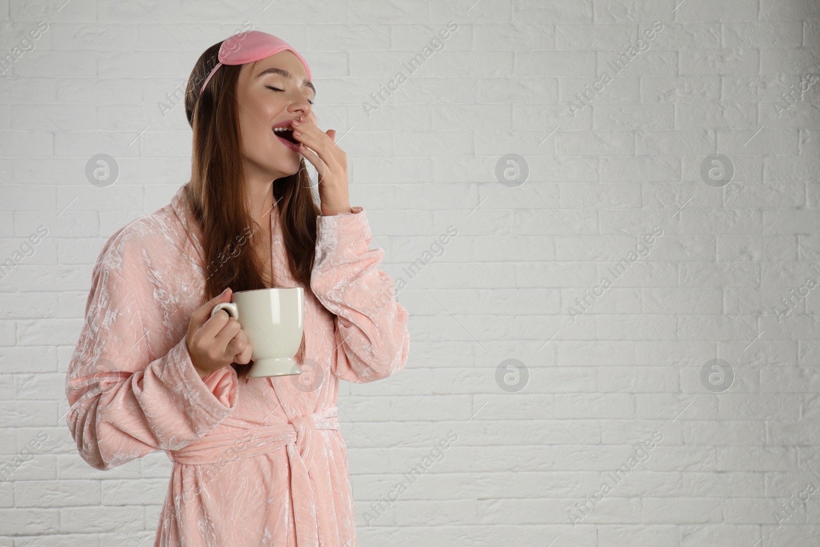 Photo of Beautiful young woman in bathrobe and eye sleeping mask yawning near white brick wall. Space for text