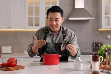 Photo of Man with pot smelling dish after cooking at countertop in kitchen