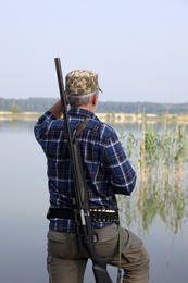 Man with hunting rifle near lake outdoors, back view