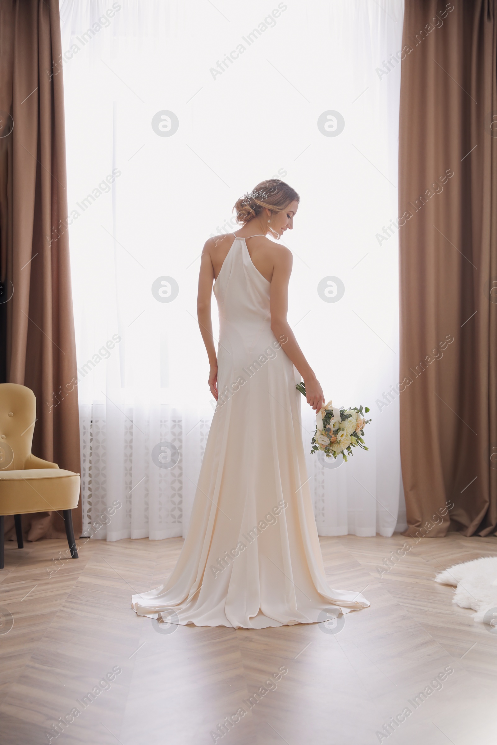 Photo of Bride in beautiful wedding dress with bouquet near window indoors, back view