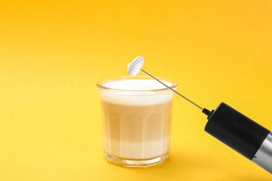 Photo of Mini mixer (milk frother) and tasty cappuccino in glass on yellow background