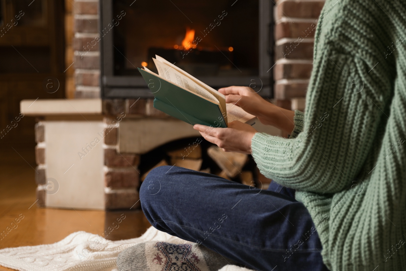 Photo of Woman reading book near burning fireplace at home, closeup