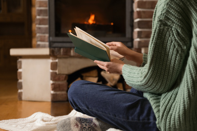 Woman reading book near burning fireplace at home, closeup