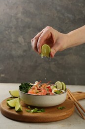 Woman squeezing lime juice into bowl of delicious ramen with shrimps at light textured table, closeup. Noodle soup