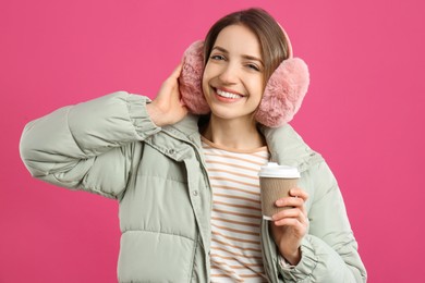 Happy woman with cup of drink wearing warm earmuffs on pink background