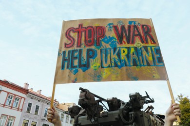 Photo of Woman holding poster Stop War Help Ukraine near broken military tank outdoors, low angle view