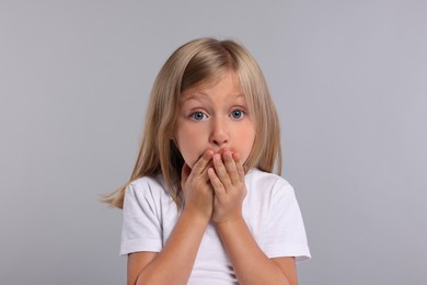 Embarrassed little girl covering mouth with hands on grey background