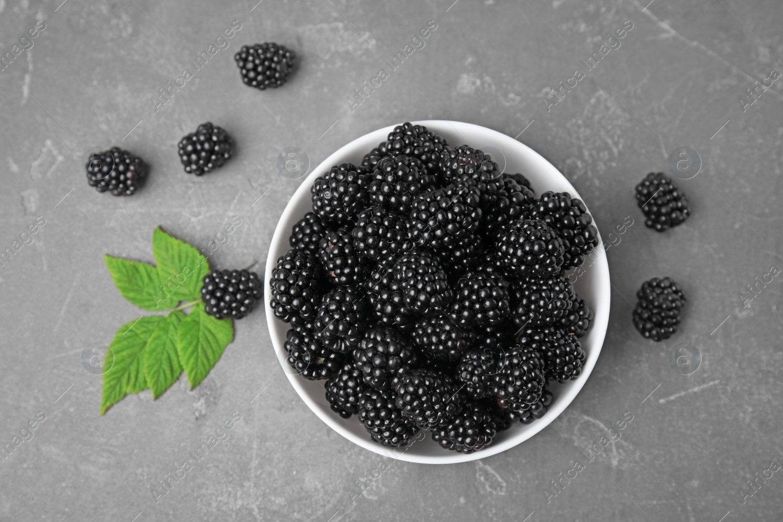 Photo of Bowl with ripe blackberries and leaves on grey table, flat lay