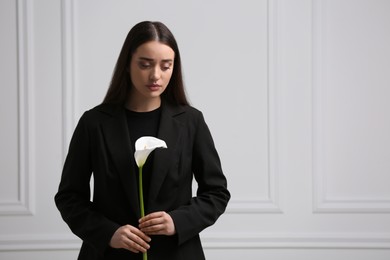 Sad woman with calla lily flower near white wall, space for text. Funeral ceremony