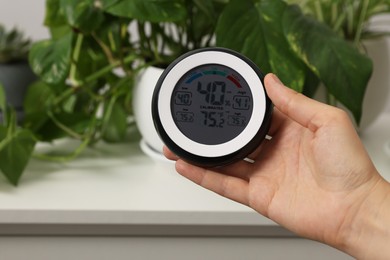 Photo of Woman holding digital hygrometer with thermometer near plants indoors, closeup