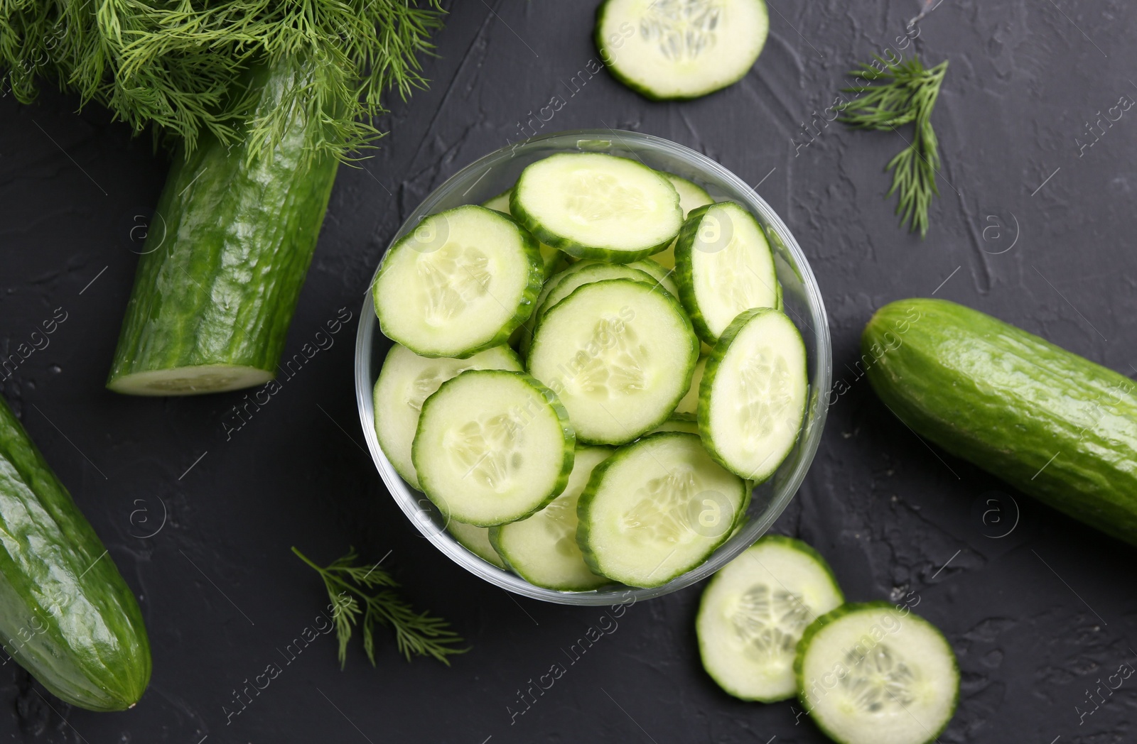 Photo of Cut cucumber in glass bowl, fresh vegetables and dill on dark gray textured table, flat lay