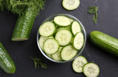 Cut cucumber in glass bowl, fresh vegetables and dill on dark gray textured table, flat lay